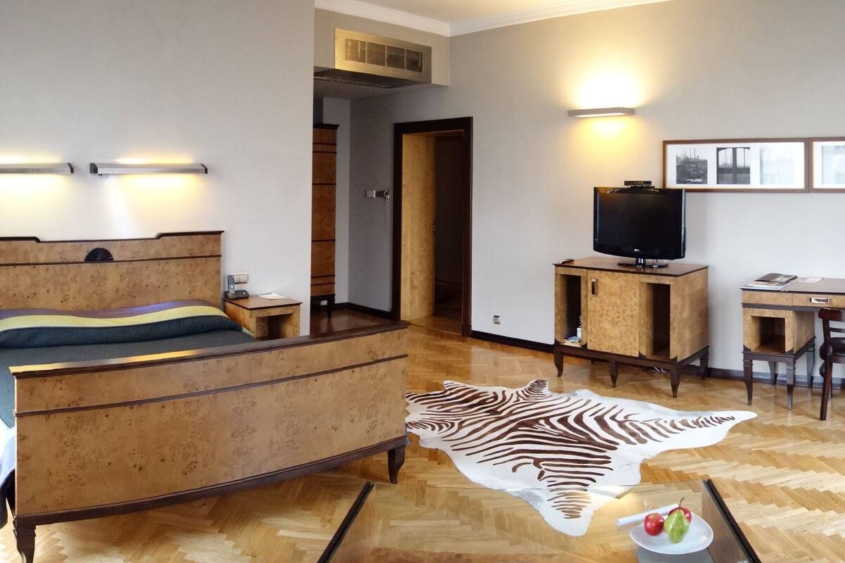 A hotel room with a wooden bed, a desk with a television, a zebra print rug, and a dining table with fruit.