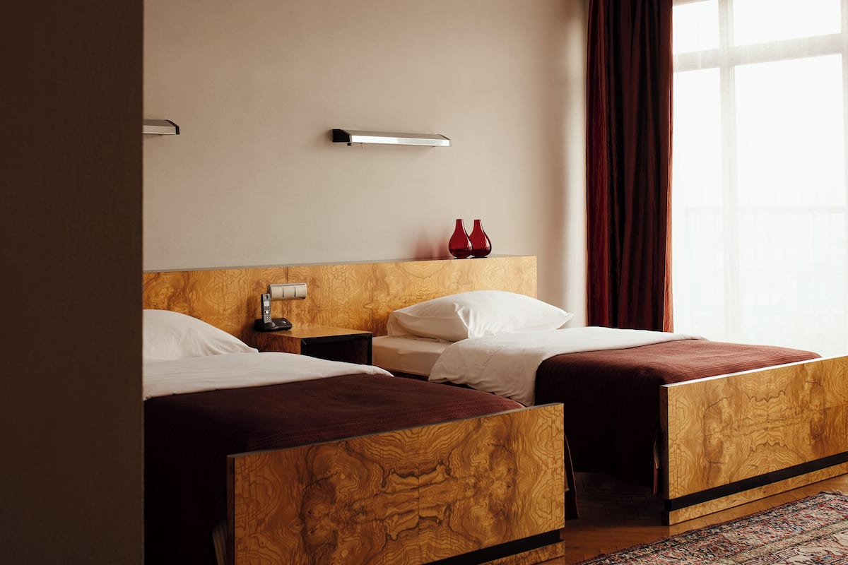 A hotel room with two wooden beds, a nightstand with a lamp, and a large window with curtains.
