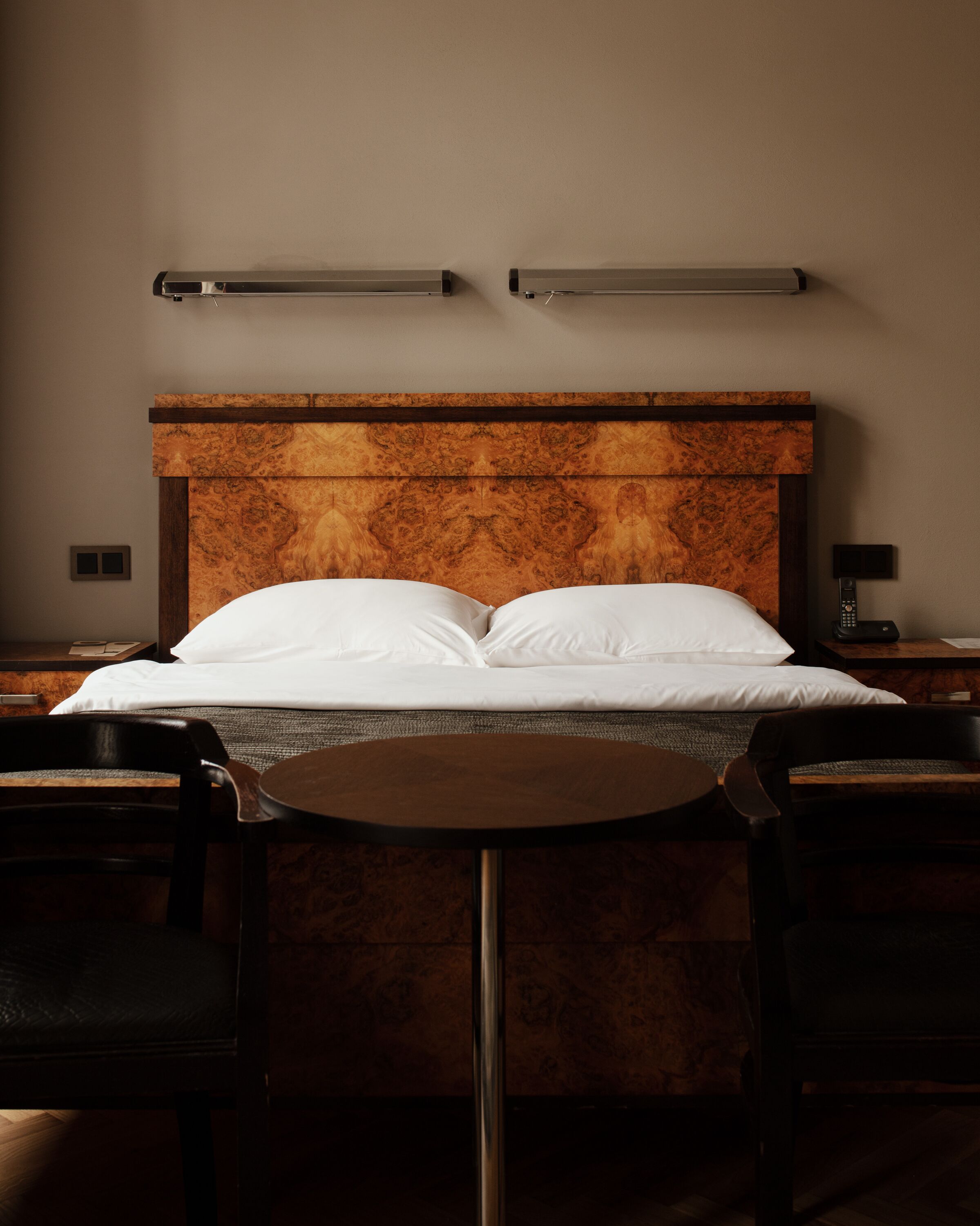 Hotelkamer met een houten hoofdbord, een ronde tafel, twee stoelen, een nachtkastje met een lamp en een aan de muur bevestigd toestel.