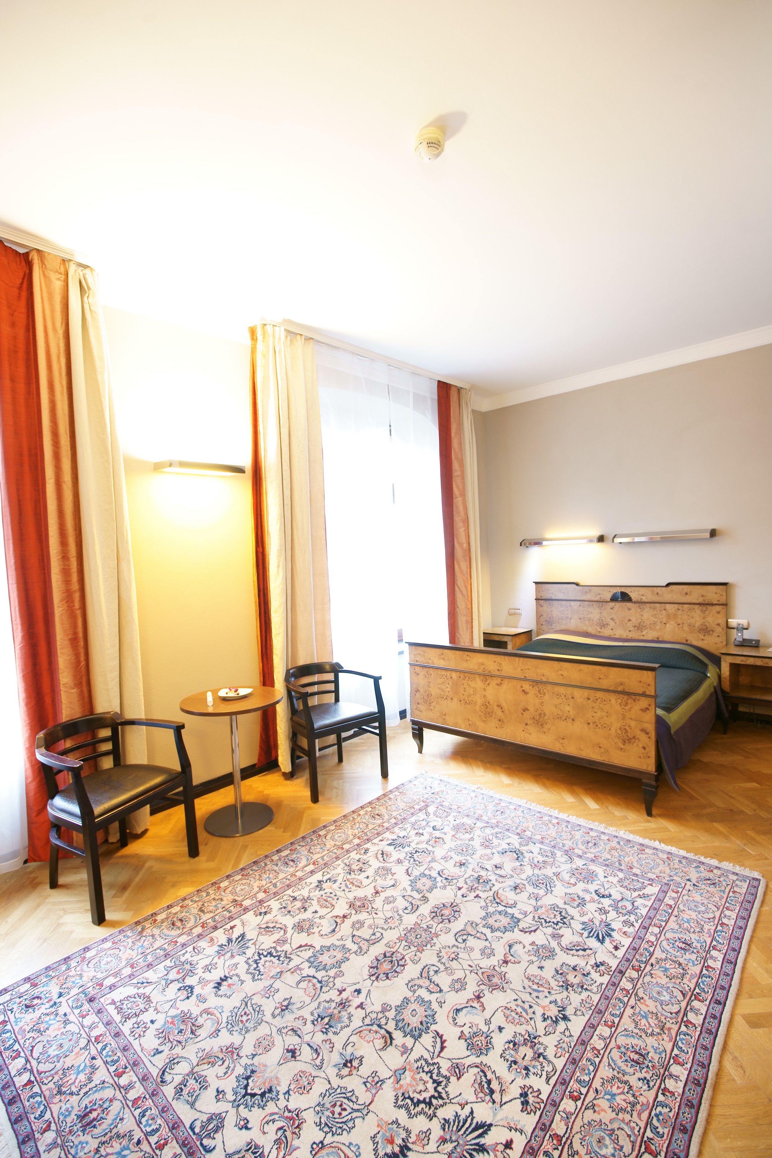 A hotel room with a bed, a desk, two chairs, a small table, and a large patterned rug.