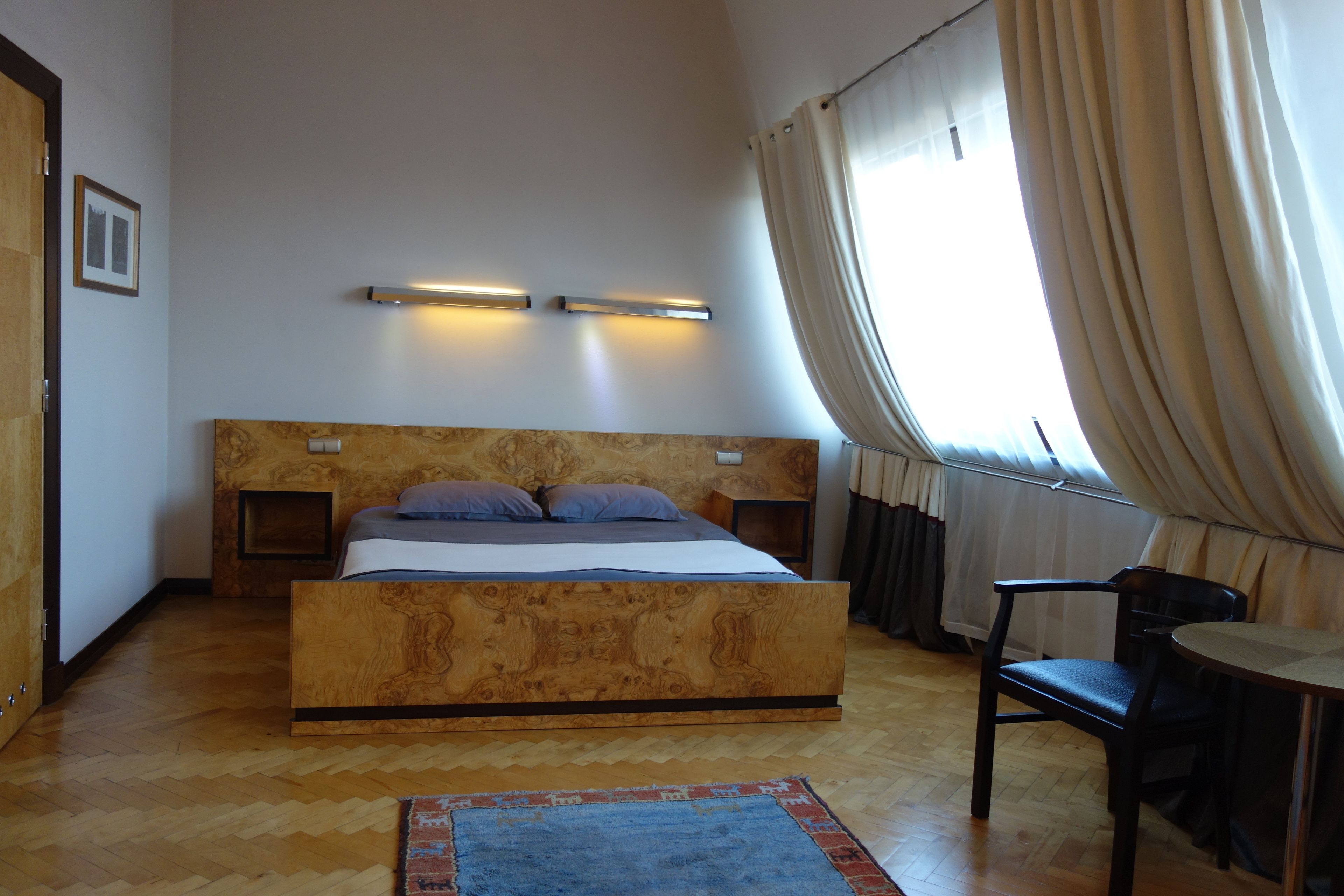 Ein Schlafzimmer mit einem hölzernen Kopfteil, einem Bett mit Kissen, einem Nachttisch, einem Sessel, einem kleinen Tisch und einem Fenster mit Vorhängen.