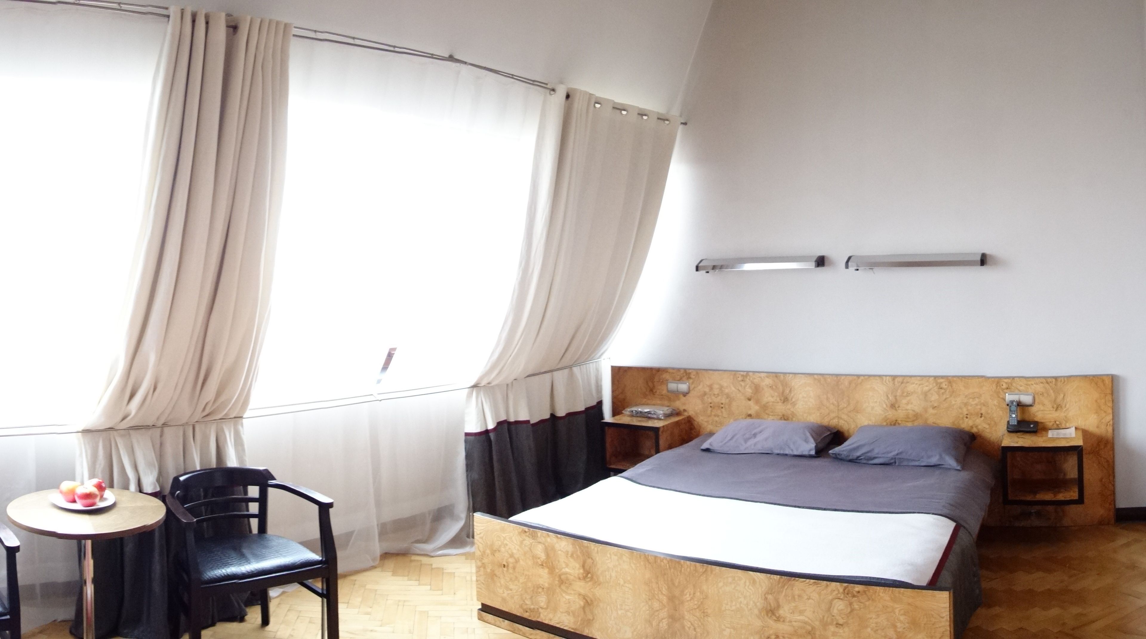 Un dormitorio con cabecera de madera, cama con ropa de cama gris, dos mesitas de noche, una pequeña mesa redonda con una silla y una ventana con cortinas.