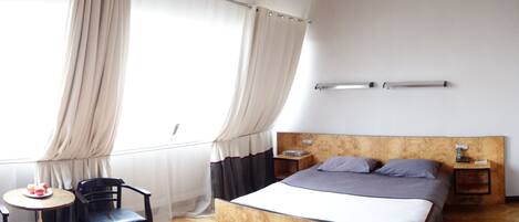 Un dormitorio con cabecera de madera, cama con ropa de cama gris, dos mesitas de noche, una pequeña mesa redonda con una silla y una ventana con cortinas.