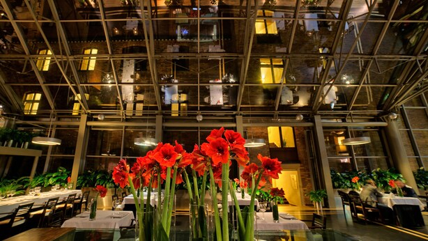 Ein modernes Restaurant mit hohen Decken und Glasverkleidungen, ausgestattet mit Esstischen und roten Blumen in Vasen.