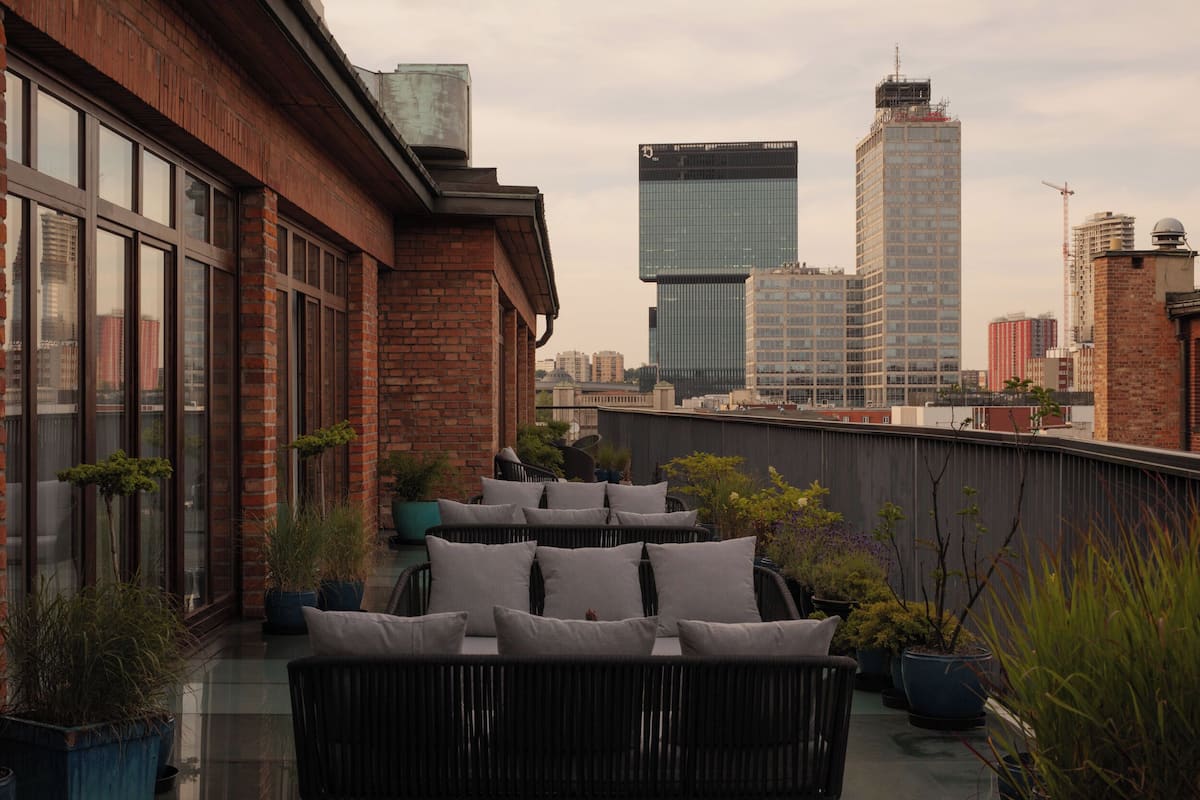 A rooftop terrace with outdoor seating, potted plants, and a city skyline in the background.