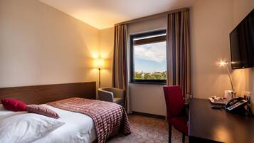 A hotel room with a bed, a desk with a phone, a chair, a lamp, and a window with curtains.