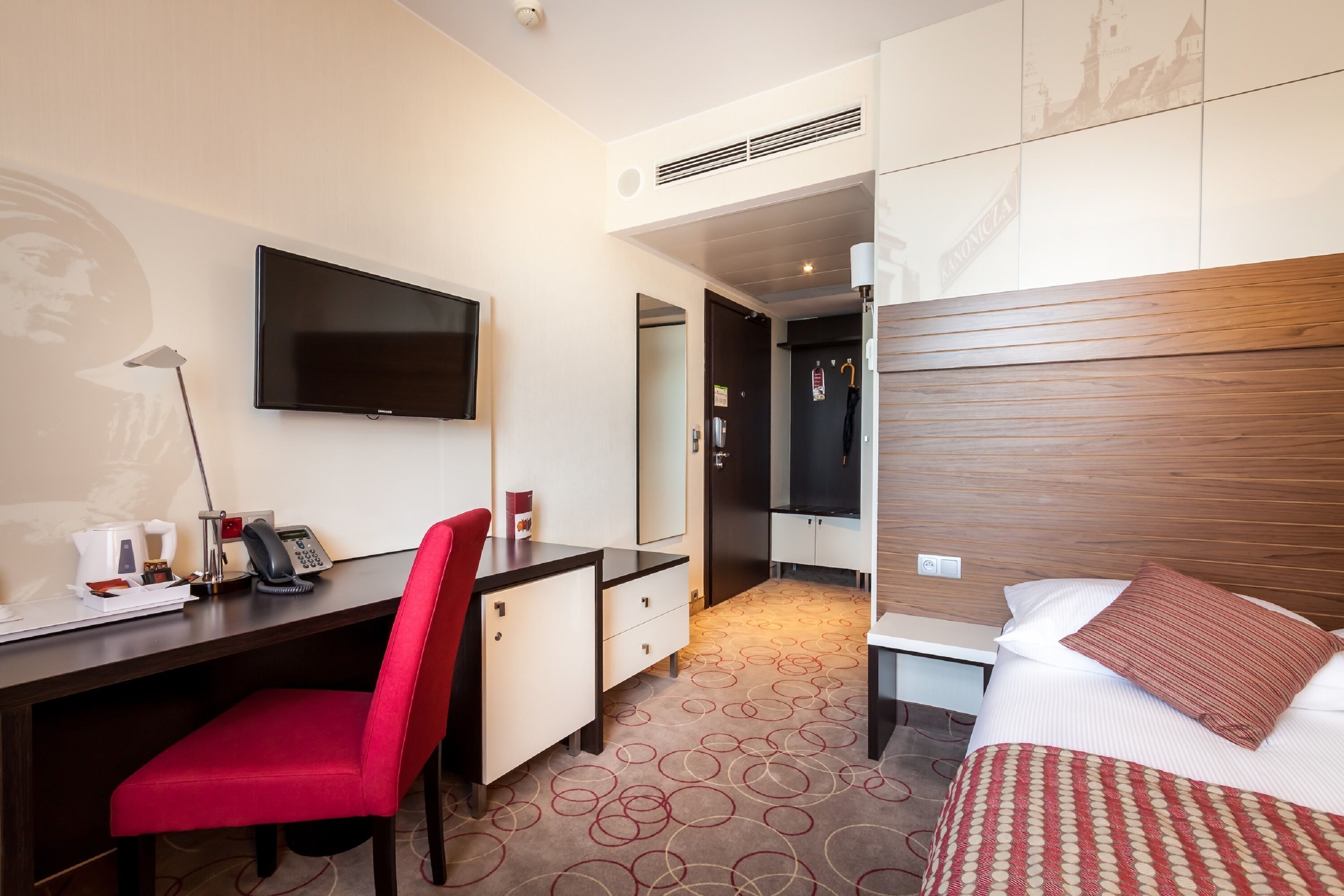 Habitación de hotel con cama, escritorio, silla, televisor y espejo.