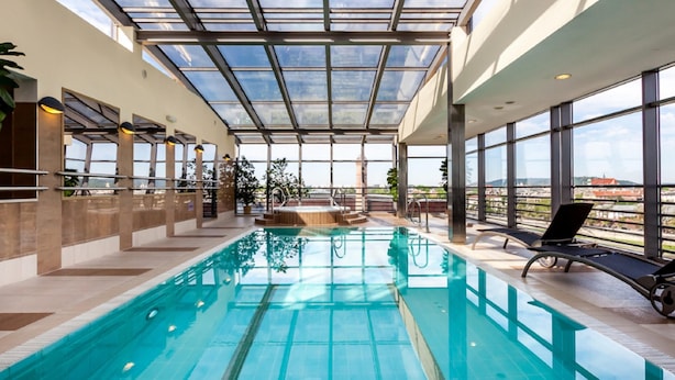 Une piscine intérieure dotée d’un toit en verre transparent, d’un intérieur baigné de lumière naturelle et offrant une vue sur le paysage urbain.