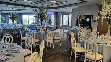 A banquet hall with round tables, white chairs, and floral centerpieces.