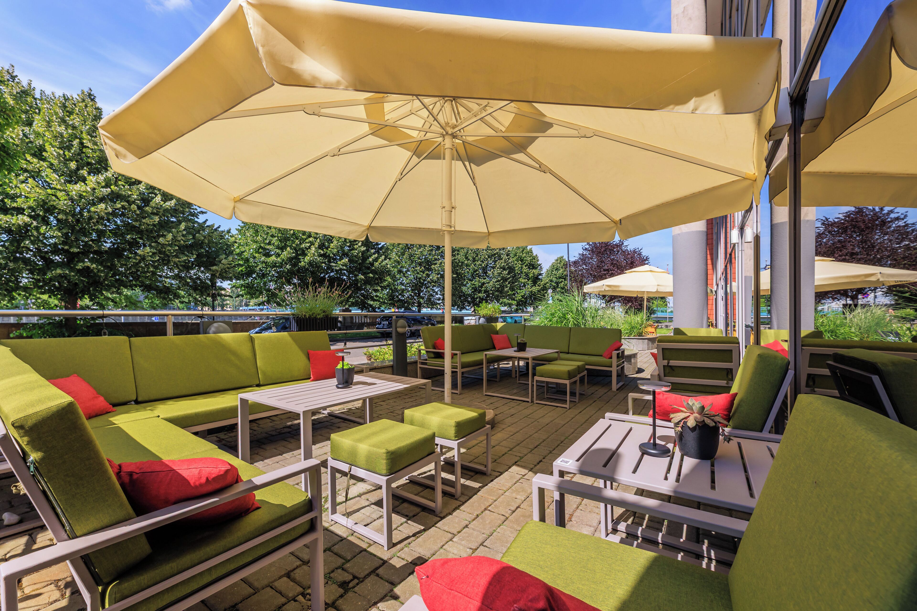 An outdoor seating area with green sofas, red cushions, and a large beige umbrella providing shade.
