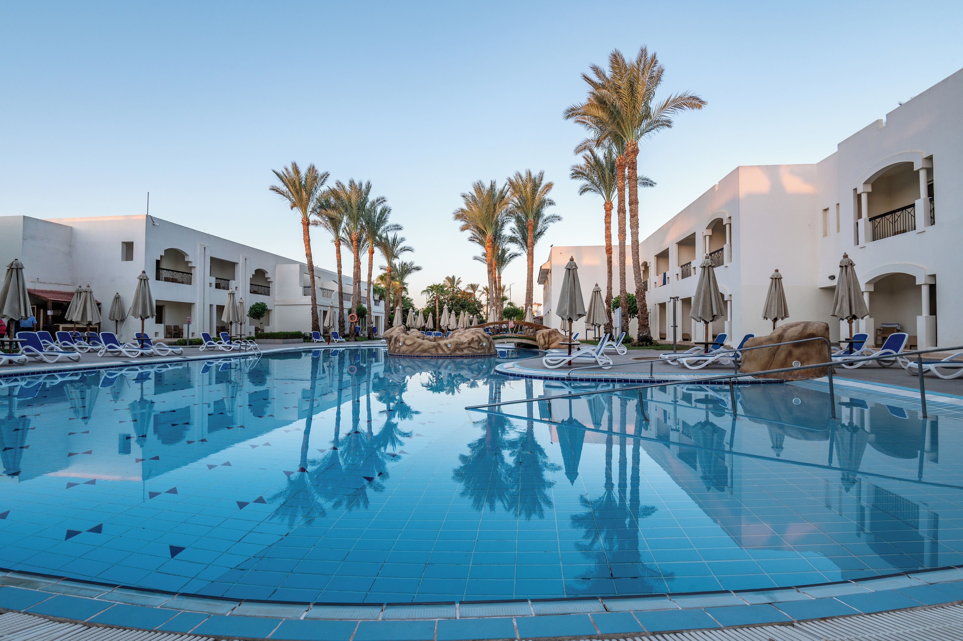 A swimming pool with lounge chairs and palm trees, surrounded by white buildings with balconies.