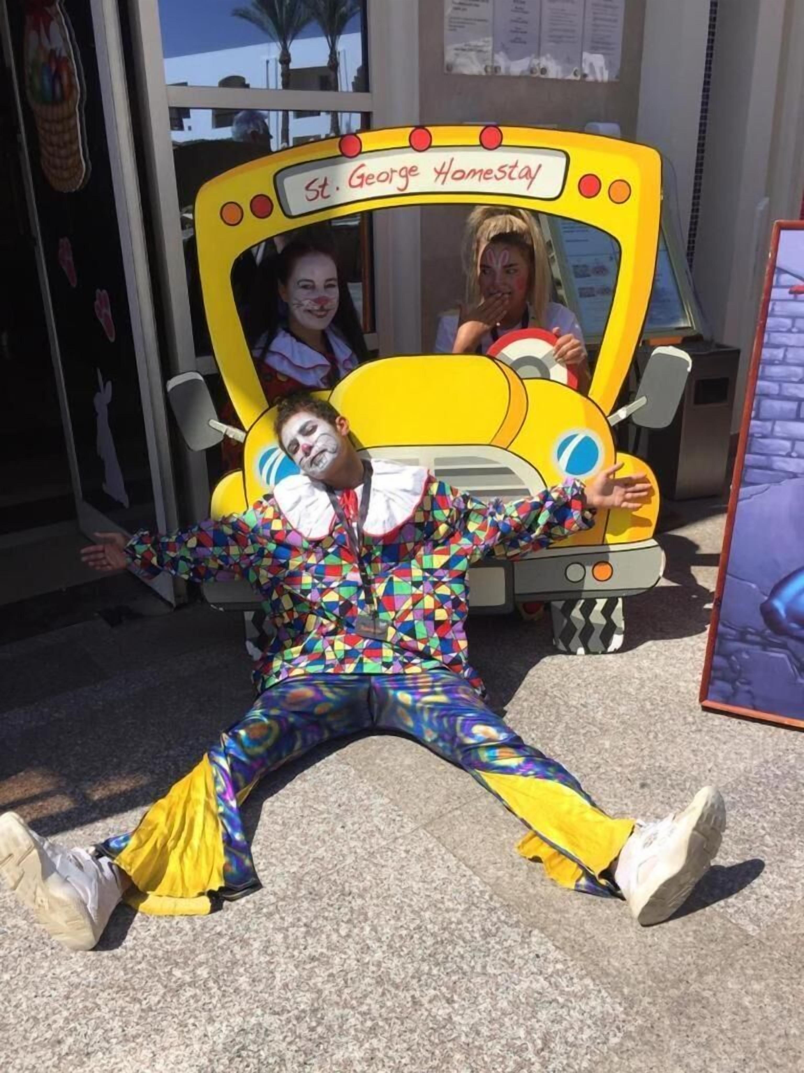 Una persona recostada en el suelo frente a un autobús amarillo con la inscripción "St. George Homestay".