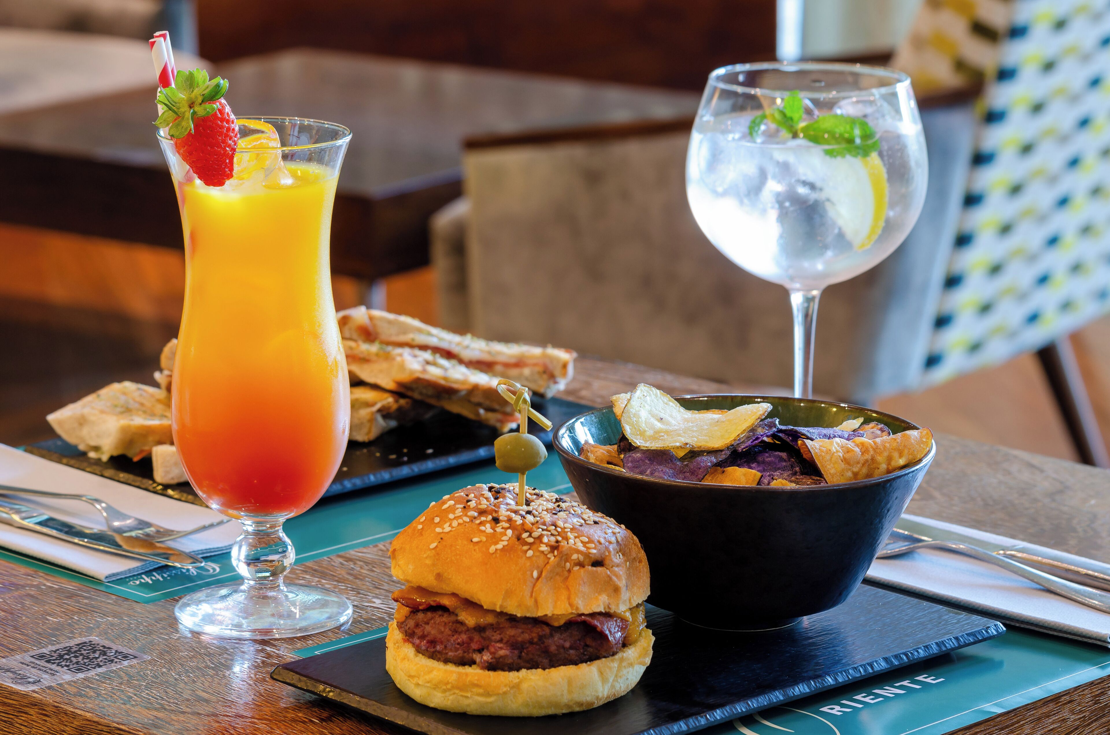 A meal with a burger, a bowl of chips, a glass of orange juice, and a glass of water with lemon and mint.