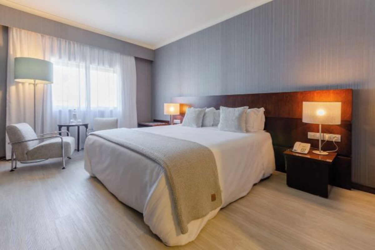 Habitación de hotel con una cama grande, dos mesitas de noche con lámparas, una silla y una ventana con cortinas.
