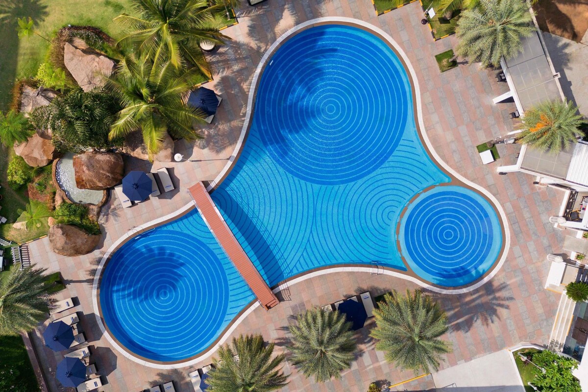 A large, kidney-shaped swimming pool with a central water slide, surrounded by palm trees and lounge chairs.