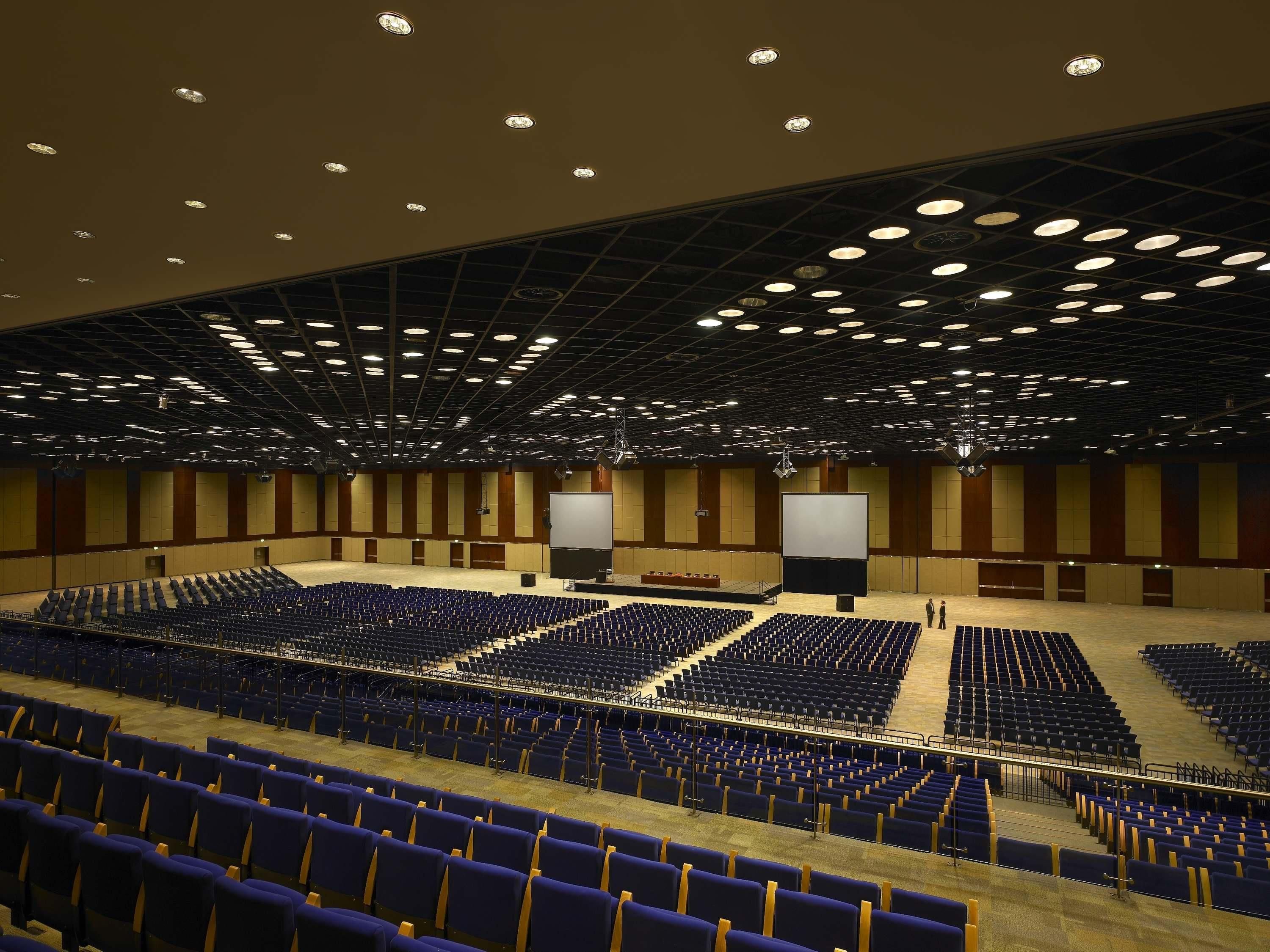 A large indoor auditorium with rows of blue seats, a stage with a large screen, and a grid of ceiling lights.