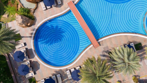 Una amplia piscina exterior de diseño circular, rodeada de sillas de descanso y sombrillas, con palmeras y rocas cerca.