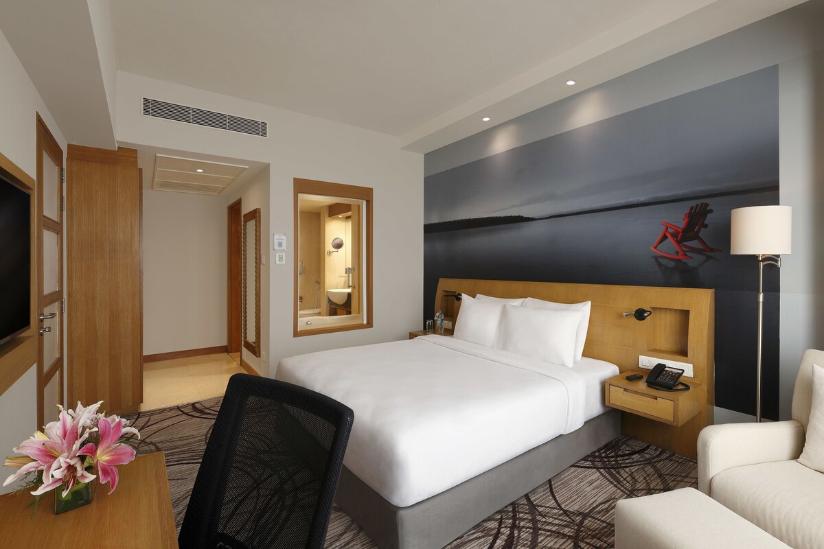 Een hotelkamer met een bed, nachtkastje, lamp, stoel, bureau, televisie en een schilderij aan de muur.