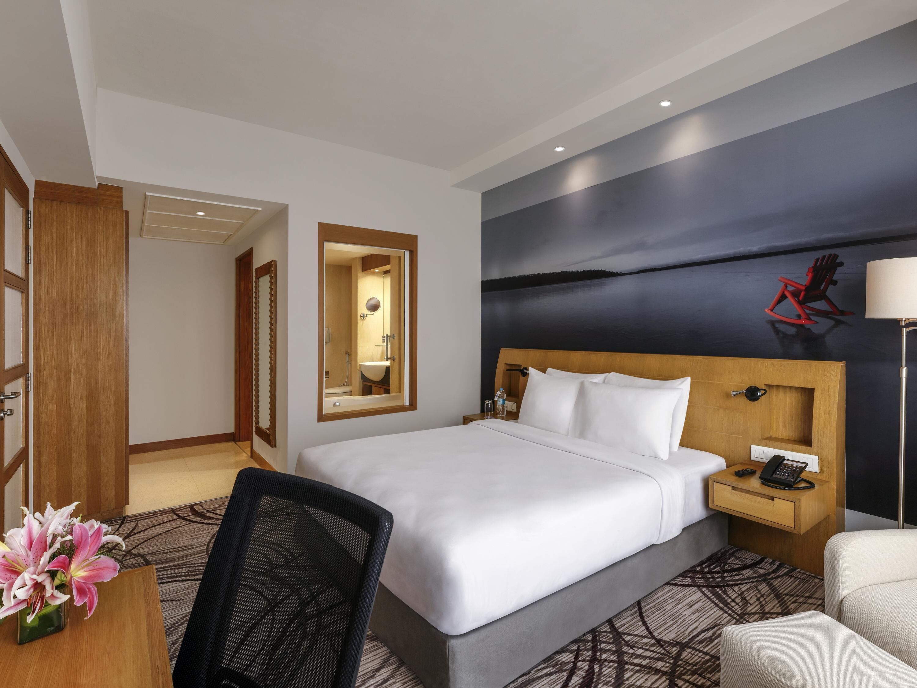 Una habitación de hotel con una cama grande, un escritorio con una silla, una mesita de noche con un teléfono y un cuadro de un paisaje en la pared.