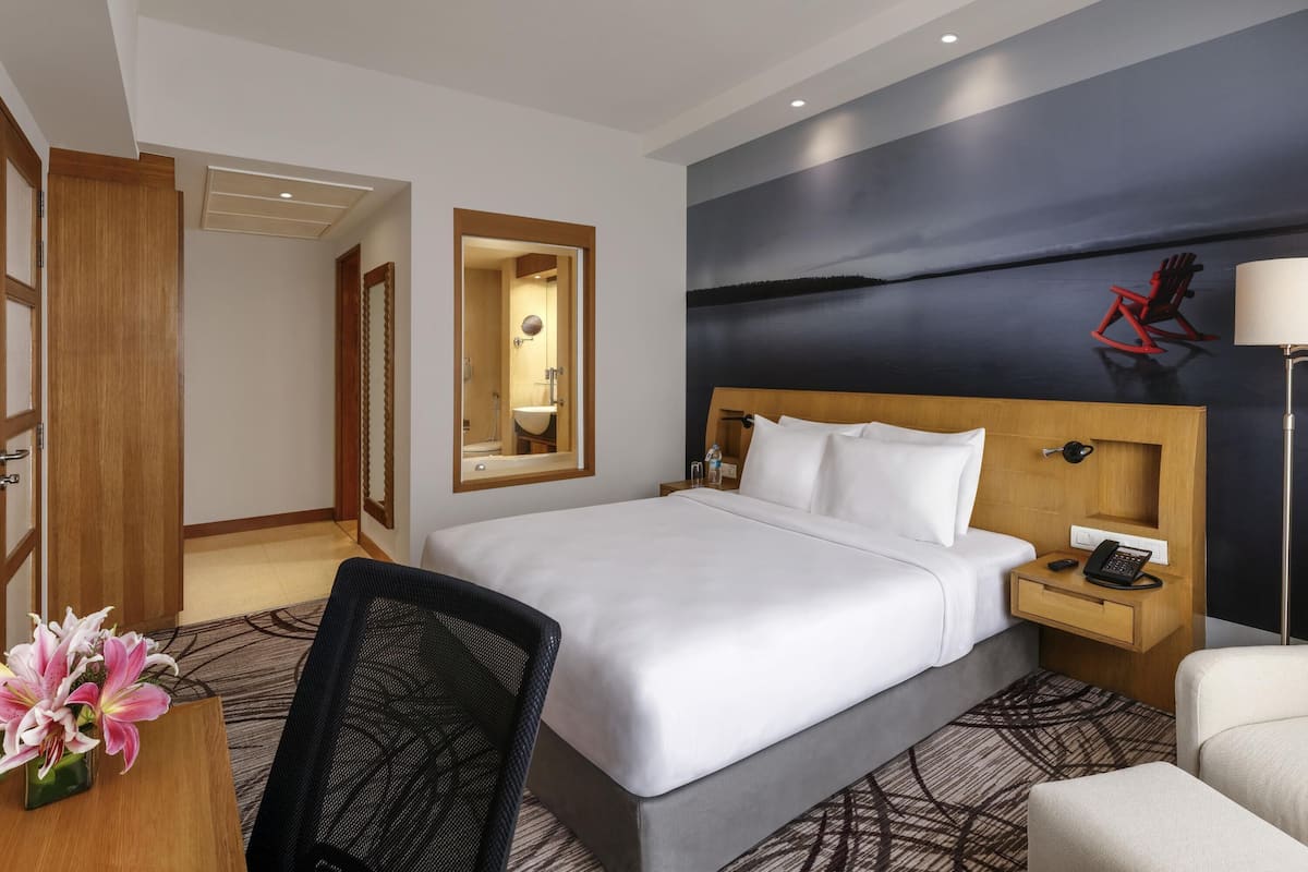 A hotel room with a large bed, a desk with a chair, a nightstand with a phone, and a painting of a landscape on the wall.