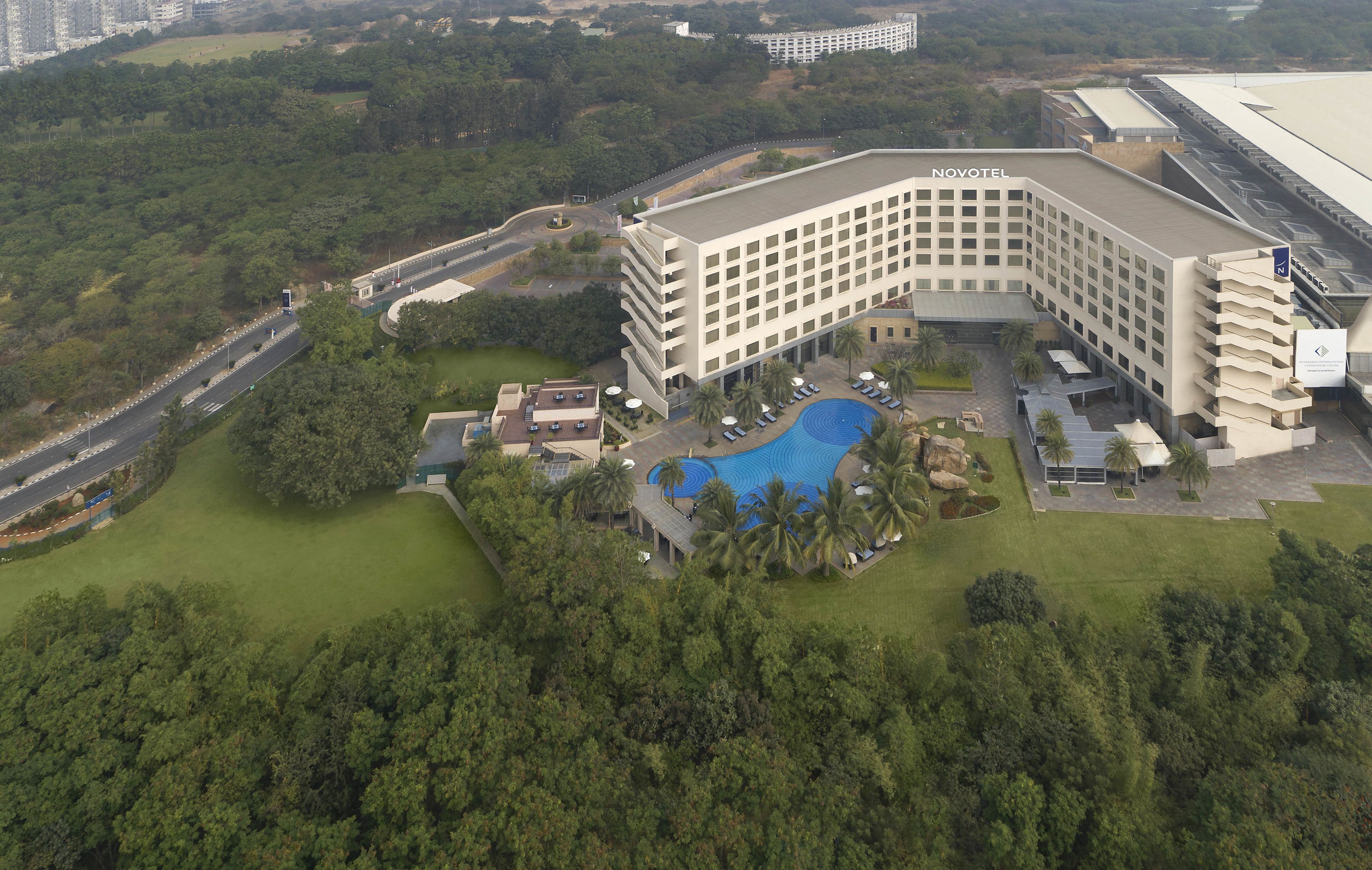 An aerial view of a hotel with a swimming pool, surrounded by greenery and a parking area.