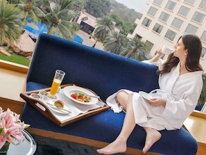 Eine Person sitzt auf einem blauen Sofa und genießt ein Frühstück mit Orangensaft, Obst und einem Croissant.