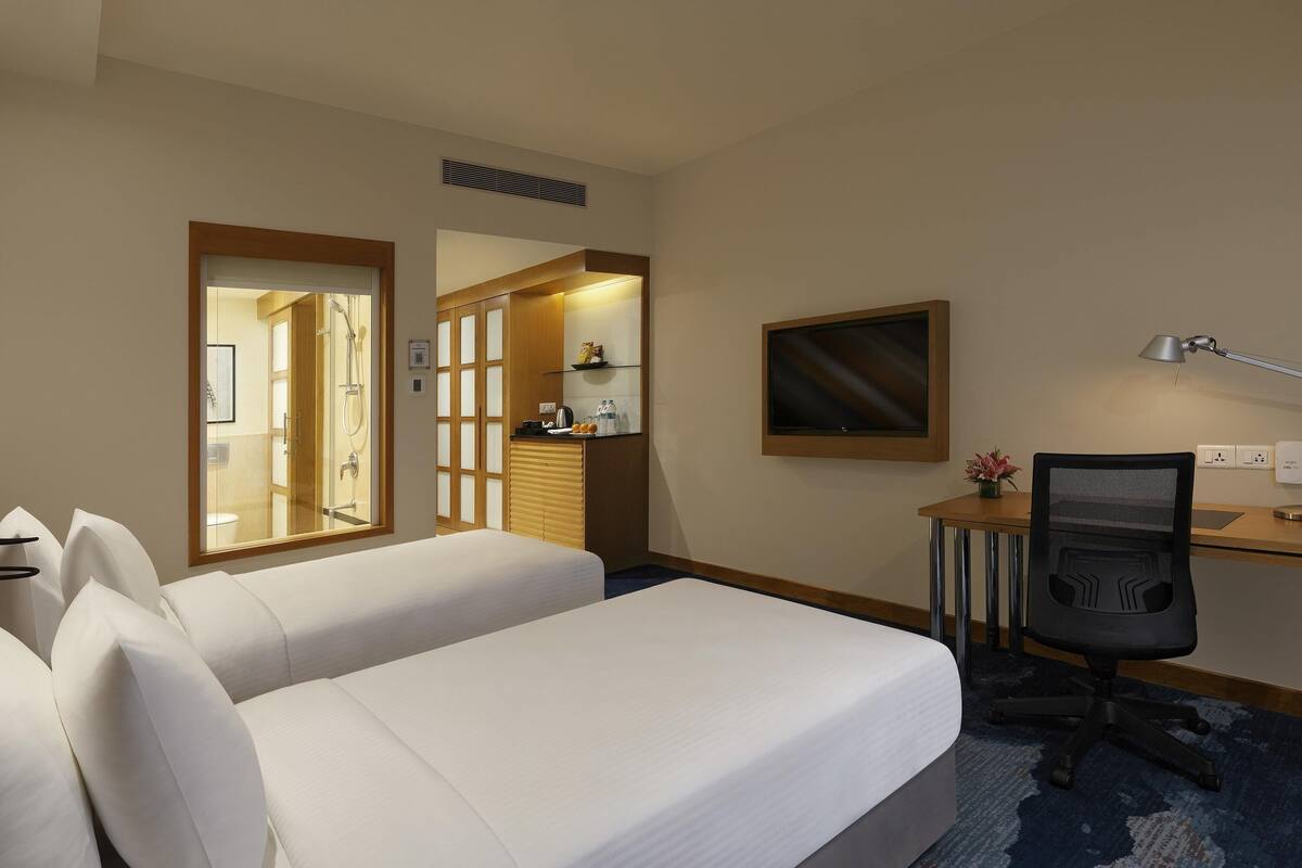 Hotellihuone, jossa on kaksi sänkyä, työpöytä, tuoli, televisio ja kylpyhuone, joka näkyy lasiovesta.
