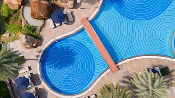 A large outdoor swimming pool with a circular design, surrounded by lounge chairs and umbrellas, with palm trees and rocks nearby.