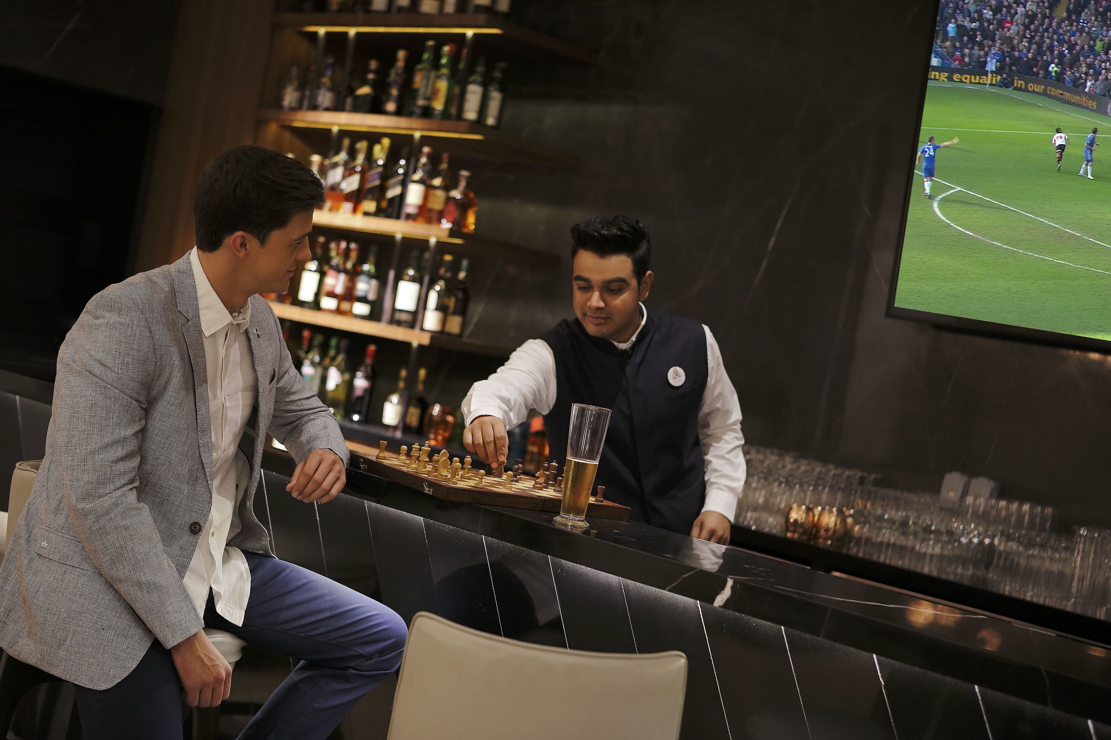 Dos hombres en un bar, con un televisor mostrando un partido de fútbol.