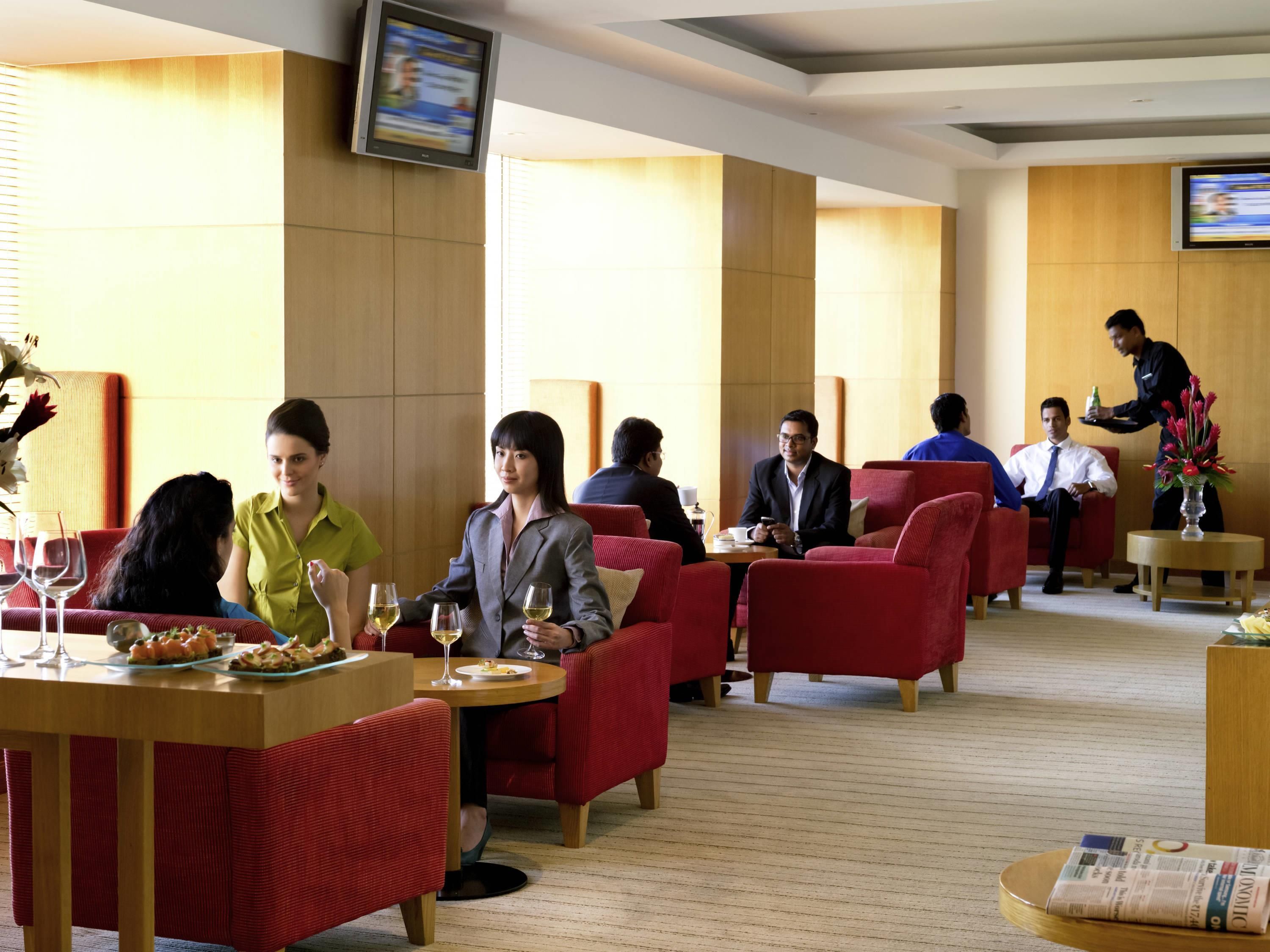 A hotel lobby with red upholstered seating, a waiter serving drinks, and a television mounted on the wall.