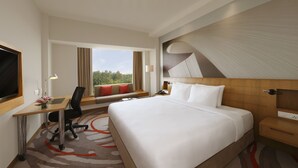 Une chambre d’hôtel avec un grand lit, un bureau avec une chaise, une télévision, une fenêtre avec des rideaux et une vue sur les arbres.