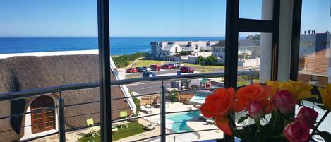 A balcony with a view of the ocean, a swimming pool, and a parking area.