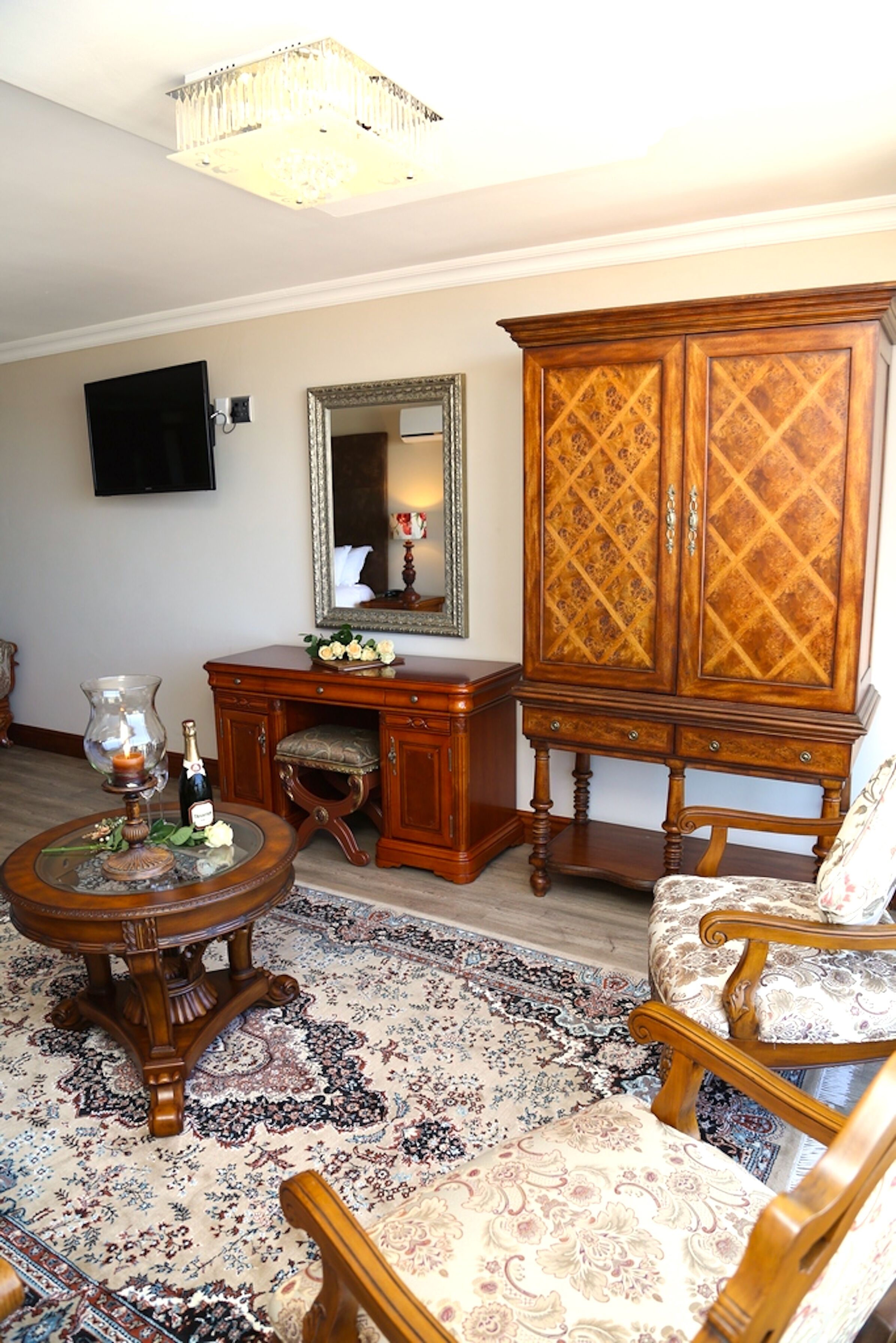 Un salon traditionnel avec un meuble en bois, un téléviseur, un miroir, une table ronde avec une bougie et un tapis à motifs.