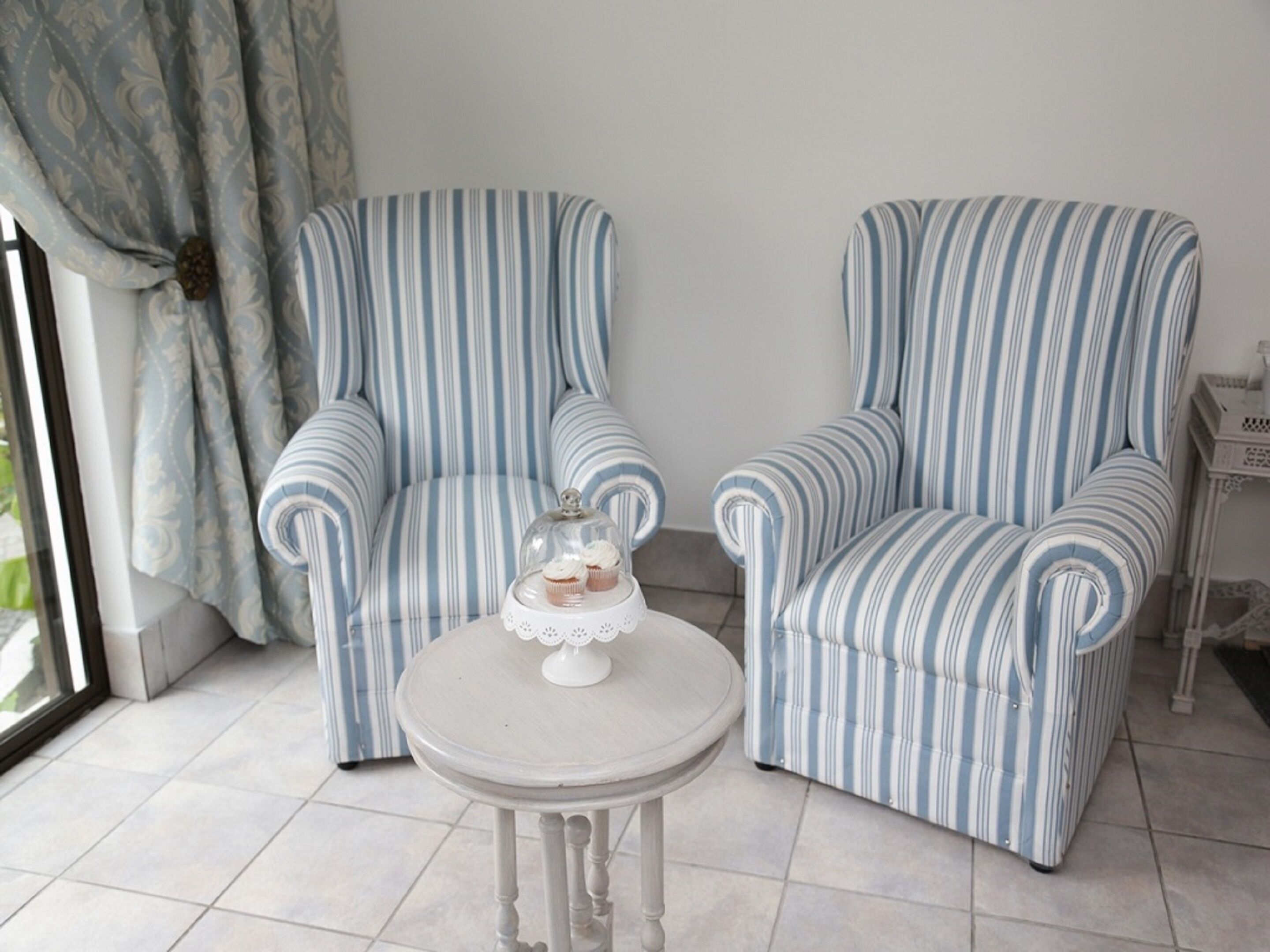 Two striped armchairs and a small round table with a glass dome centerpiece in a room with tiled flooring and a window with curtains.