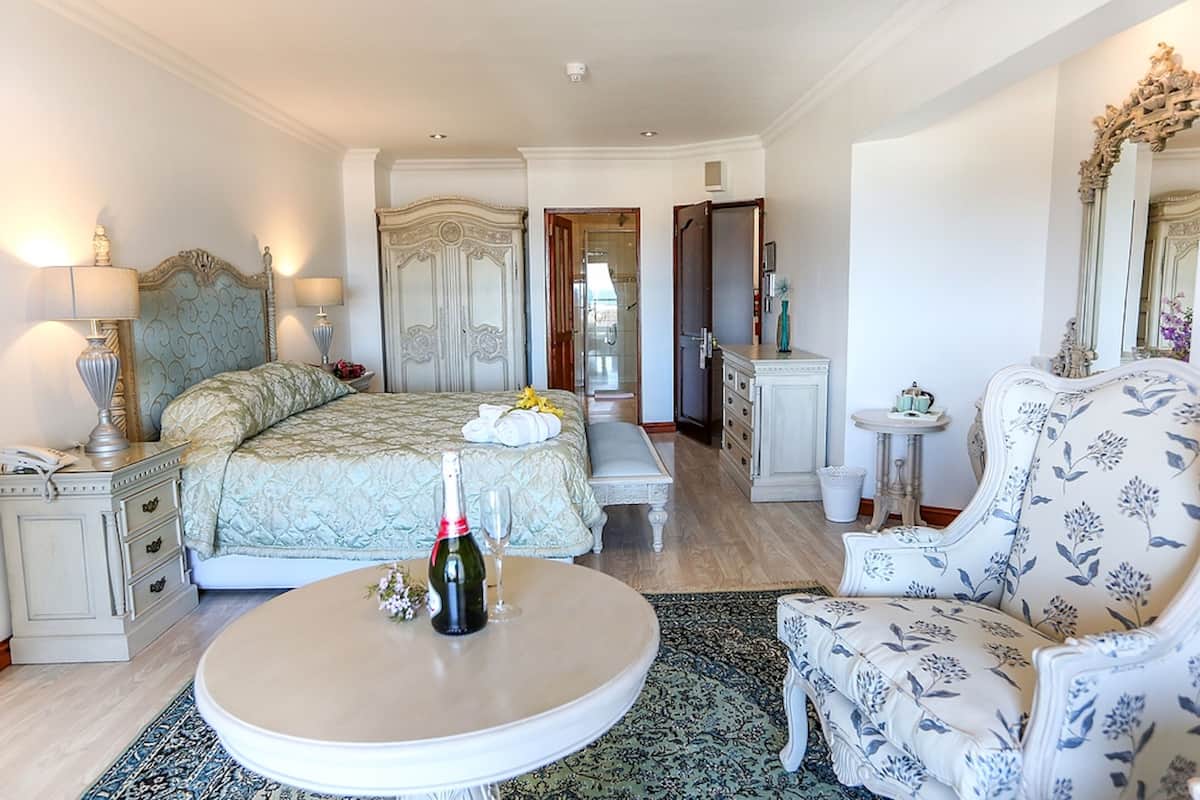 Une chambre d’hôtel avec un lit, une table basse ronde sur laquelle se trouvent une bouteille et un verre, une chaise recouverte d’un tissu à motifs floraux, une commode et un miroir avec un cadre orné.