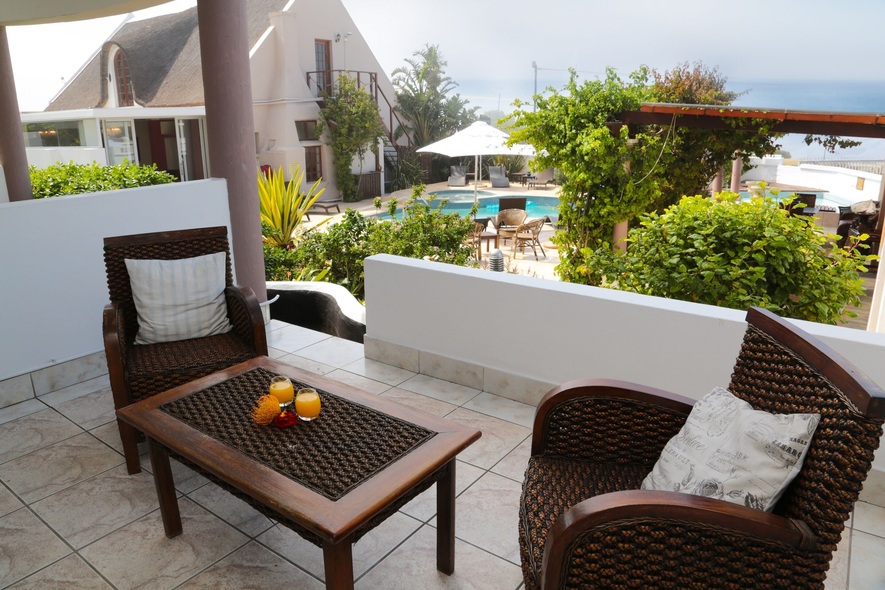 A balcony with wicker furniture, a table with a glass of orange juice, and a view of a swimming pool and greenery.