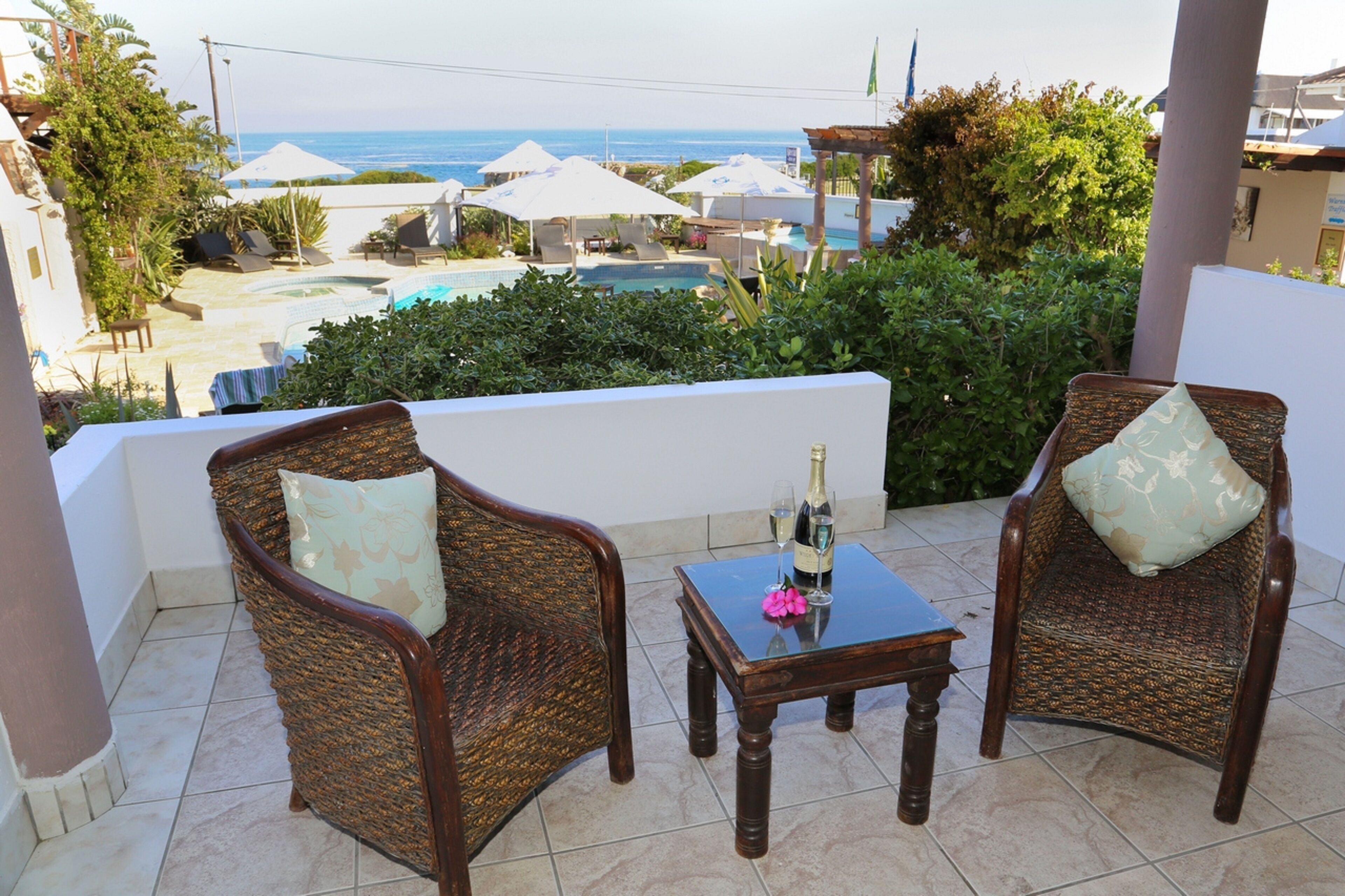 A balcony with wicker chairs, a table with a bottle and glass, and a view of the sea and pool area.
