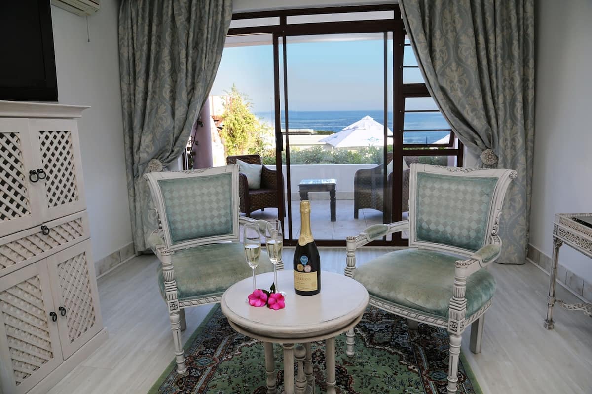 Une pièce comprenant deux fauteuils rembourrés, une petite table ronde avec une bouteille de champagne et deux verres, et une vue sur la mer grâce à de grandes fenêtres.
