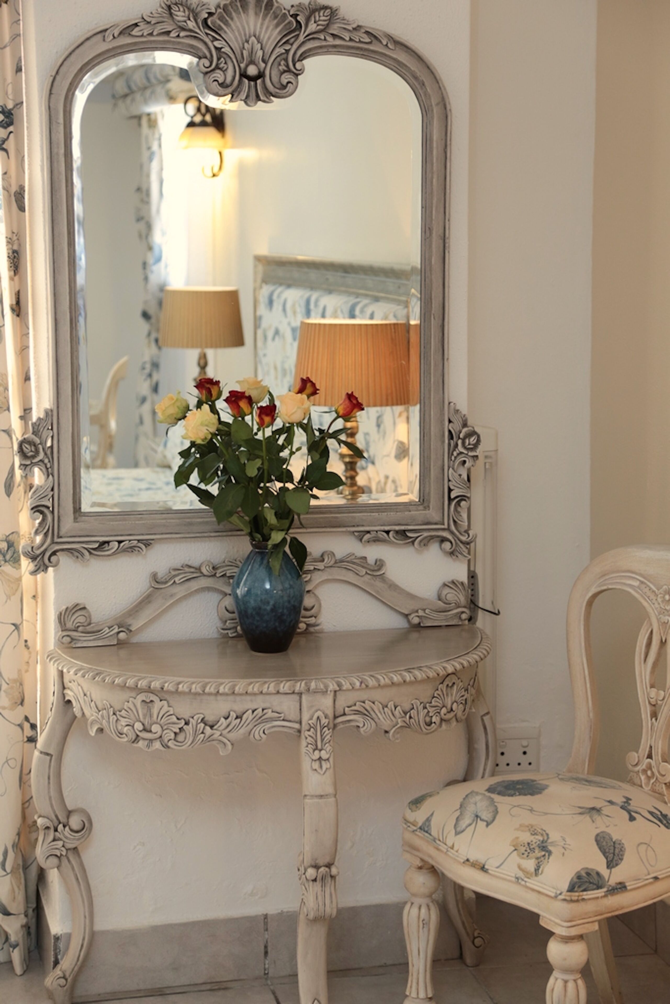 A vintage-style vanity with a mirror, a blue vase with flowers, and a chair with floral upholstery.