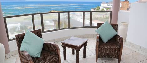A balcony with wicker chairs and a small table, overlooking a coastal view.