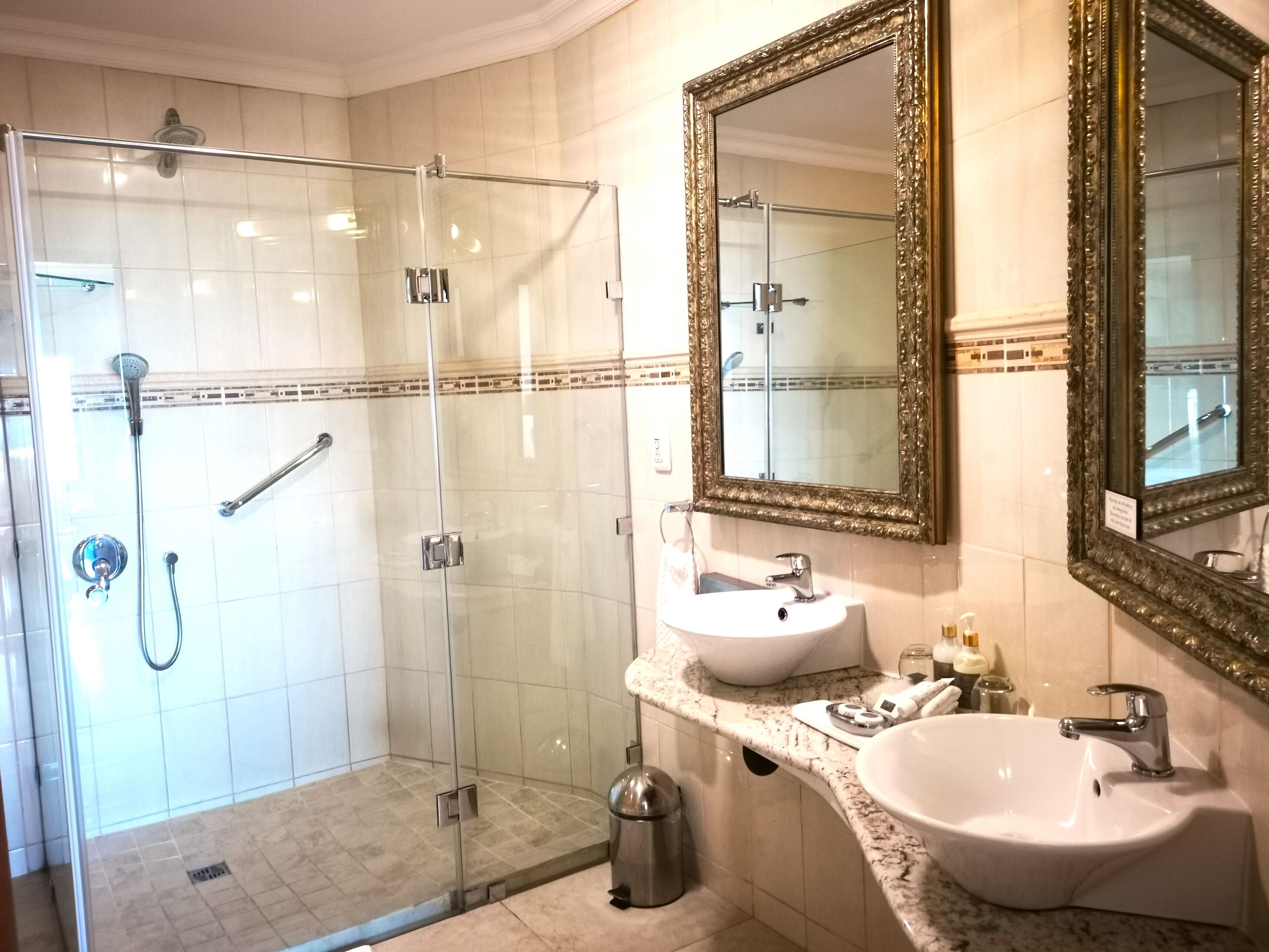 Een moderne badkamer met een douchecabine van glas, een witte wastafel en twee grote spiegels met sierlijke lijsten.