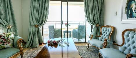 A room with a wooden coffee table, two upholstered armchairs, a sofa, and a balcony with a view.