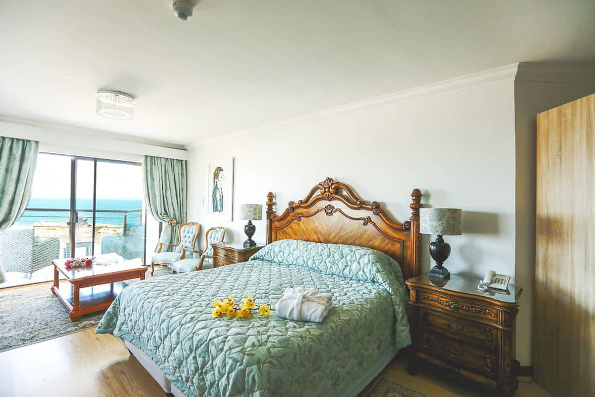 Une chambre spacieuse avec un grand lit, une tête de lit en bois, une table de chevet avec une lampe et une vue sur la mer par une grande fenêtre.