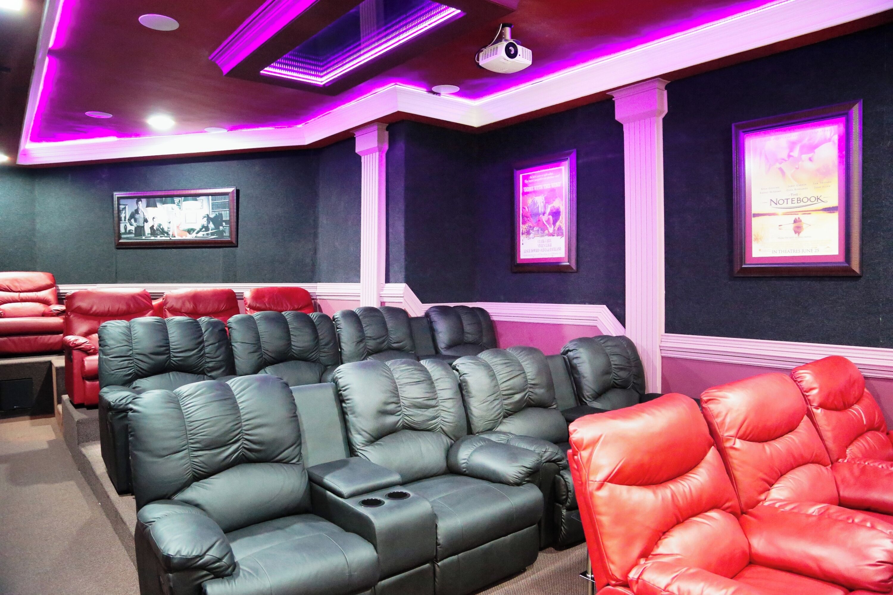 A theater with rows of black and red reclining seats, a ceiling projector, and framed pictures on the walls.