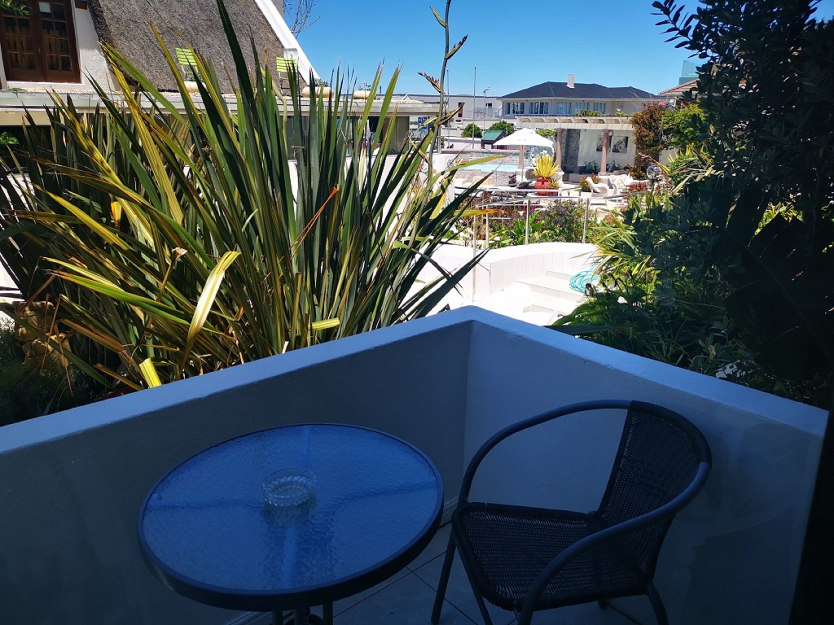 Un balcón con mesa y silla de vidrio, con vistas a un área de piscina con tumbonas y sombrillas.