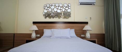 A hotel room with a large bed, two bedside tables with lamps, a wall-mounted air conditioning unit, and a decorative mirror arrangement.