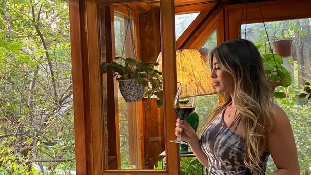 A woman holding a wine glass in a room with wooden beams, a table with wine bottles and glasses, and a view of greenery outside. - Casa Glebinias Hotel Jardin (Chacras de Coria)