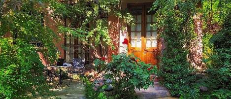 A rustic garden with a wooden bench and a table, surrounded by lush greenery and a stone pathway.