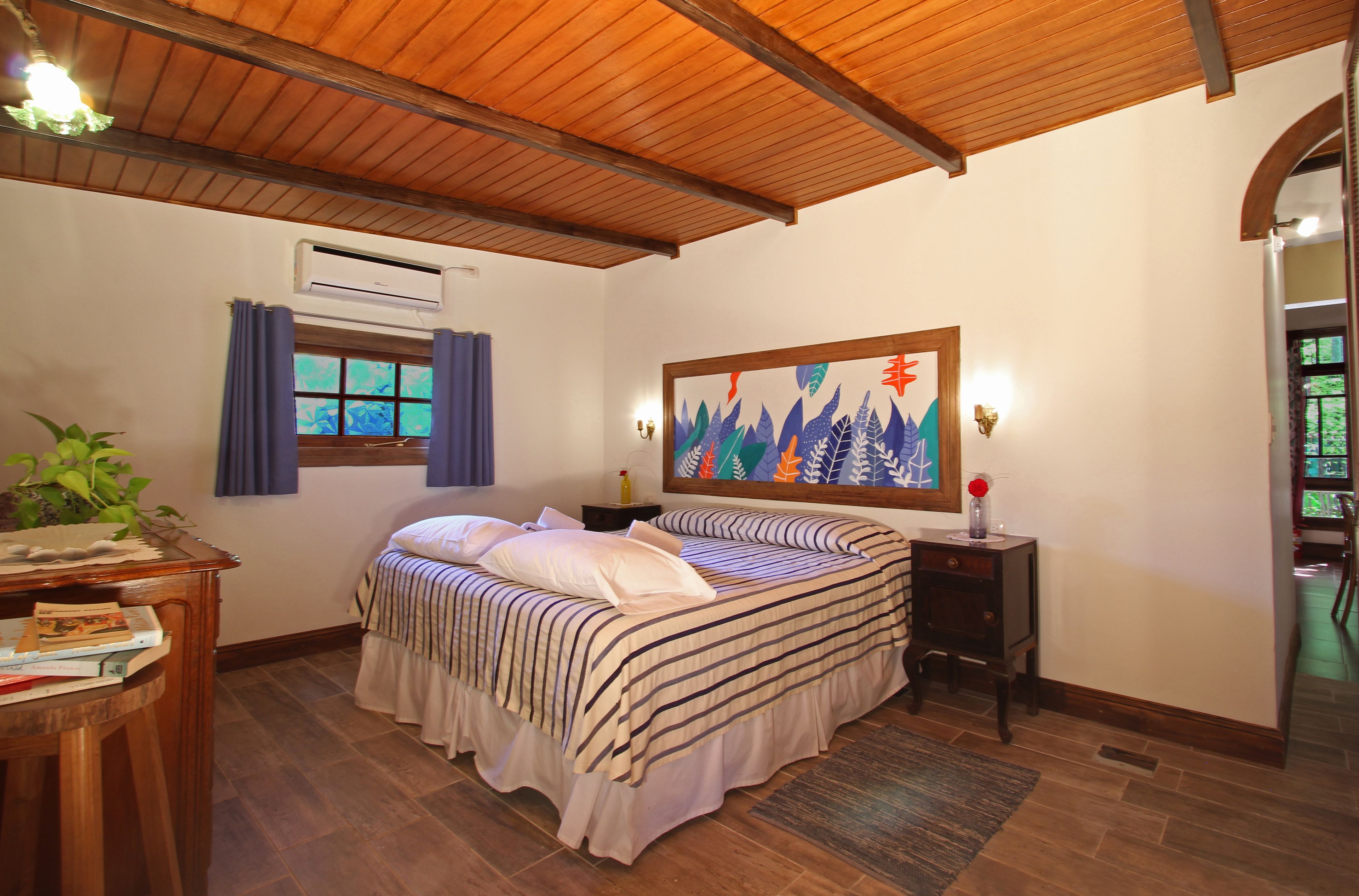 A bedroom with a wooden ceiling, a bed with striped bedding, a nightstand with a lamp, a wall-mounted air conditioner, and a window with curtains.