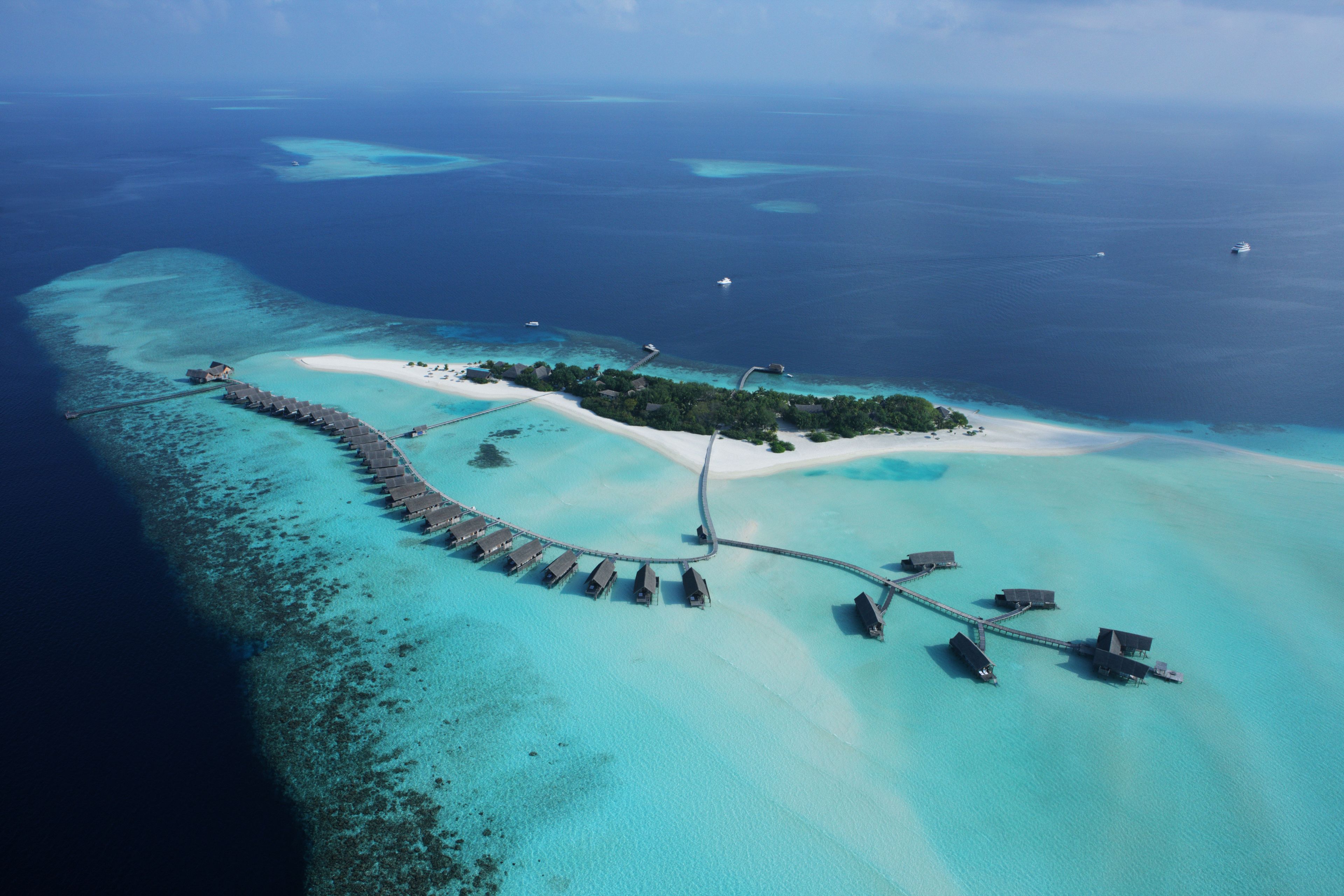 An aerial view of a tropical island resort with a long pier connecting to the mainland, surrounded by clear turquoise waters and small islets.